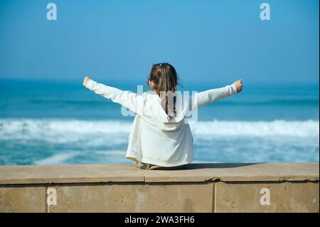 Rückansicht eines kleinen Mädchens, das ihre Arme zur Seite ausstreckt, am Rand des Atlantischen Ozeans sitzt und den faszinierenden Blick auf das wunderschöne Fu genießt Stockfoto