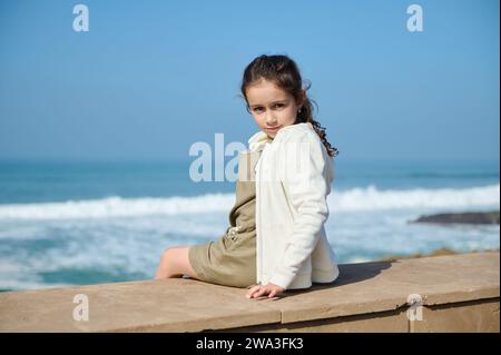 Entzückendes weißes kleines Mädchen, das auf die Kamera blickt und allein am Meer vor dem Hintergrund des Atlantischen Ozeans sitzt. Menschen. Lifestyle. Leisur Stockfoto