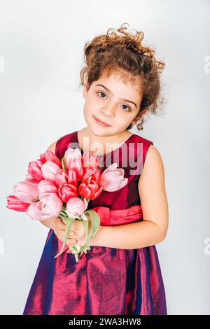 Studio-Aufnahme eines 5-jährigen Mädchens mit lockigen Haaren, mit Partykleid und einem kleinen Strauß frischer rosafarbener Tulpen Stockfoto
