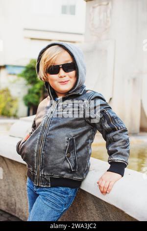 Außenporträt eines entzückenden kleinen blonden Jungen mit schwarzer Lederjacke und Sonnenbrille Stockfoto