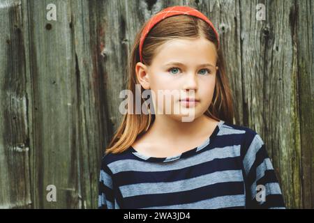 Außenporträt eines süßen kleinen 8-9-jährigen Mädchens mit braunen Haaren, mit Streifenspitze, stehend vor altem hölzernem Hintergrund Stockfoto