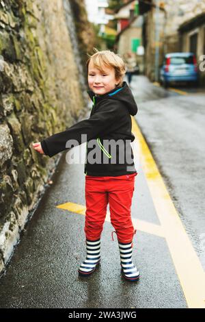 Kleiner Junge, der im Frühjahr draußen spielt, neben der Straße läuft, schwarze, wasserdichte Jacke, rote Hose und Regenstiefel trägt Stockfoto