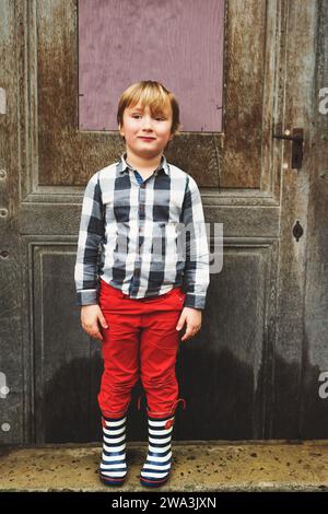 Der kleine Junge posierte im Frühjahr draußen, stand neben dem alten Hintergrund, trug ein graues kariertes Hemd, eine rote Hose und Regenstiefel Stockfoto