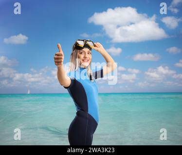 Eine junge Frau im Meer, die einen Taucheranzug und eine Maske trägt und die Daumen nach oben bewegt Stockfoto