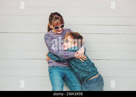 Gruppe von zwei lustigen Kindern, die draußen zusammen spielen, kleiner Junge und Mädchen posieren vor weißem hölzernem Hintergrund, Bruder und Schwester tragen passende Zugkraft Stockfoto