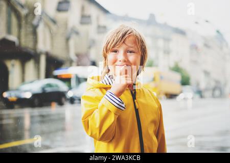 Süßer kleiner Junge, der unter dem Regen in einer Stadt spielt, in einem hellgelben Regenmantel mit Kapuze. Bild aufgenommen auf dem Saint-Francois-Platz, Lausanne, Schweiz Stockfoto