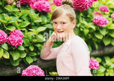 Außenporträt eines hübschen kleinen Mädchens, das mit wunderschönen Hortensie-Blumen im Sommergarten spielt, ein Kleid im Ballerina-Stil trägt, Haarkochen Stockfoto