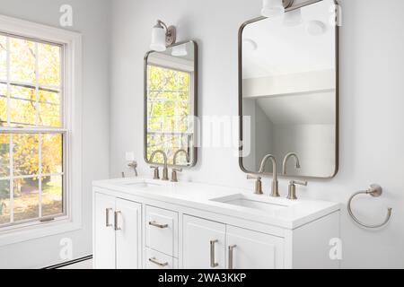 Ein Badezimmer mit weißem Schrank, bronzenen Armaturen und Spiegeln und bunten Bäumen aus den Fenstern. Stockfoto