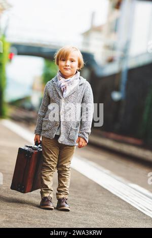 Entzückender kleiner Junge auf einem Bahnhof, der mit Koffer auf den Zug wartet Stockfoto