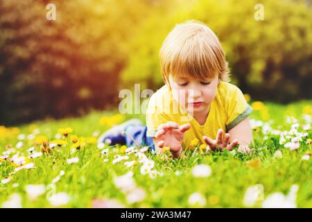Sonniges Sommerporträt eines jungen hübschen kleinen Jungen, der an einem schönen warmen Tag draußen spielt, auf hellgrünem Gras liegend, mit gelbem T-Shirt Stockfoto