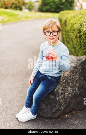 Outdoor-Porträt eines niedlichen kleinen Jungen mit Brille, hellblauem Pullover, Jeans und weißen Körben, der einen Drink mit einem Strohhalm hält Stockfoto