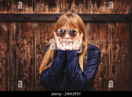 Outdoor-Modeporträt eines lustigen 9-10-jährigen Mädchens mit Sonnenbrille und blauer Lederjacke Stockfoto