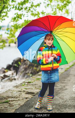 Außenporträt eines hübschen kleinen Mädchens mit großem bunten Regenschirm, Regenjacke und Stiefel, Wetterkonzept, Mode für Kinder Stockfoto