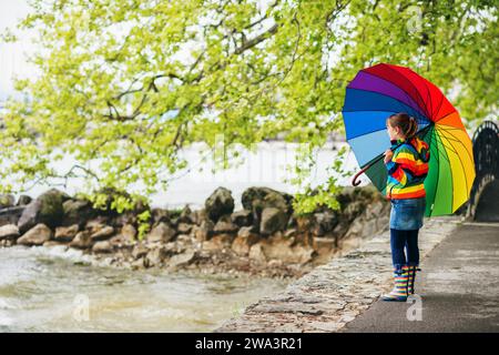 Außenporträt eines hübschen kleinen Mädchens mit großem bunten Regenschirm, Regenjacke und Stiefel, Wetterkonzept, Mode für Kinder Stockfoto