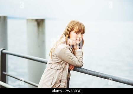 Outdoor-Porträt eines entzückenden 9- bis 10-jährigen Mädchens mit modernem beigefarbenem Trenchcoat, das an einem kalten Tag in einem Hafen posiert Stockfoto