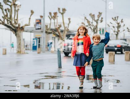 Zwei lustige Kinder, kleiner Bruder und Schwester spielen zusammen in einer Stadt unter dem Regen im Frühjahr. Kinder mit Regenjacken und Stiefeln Stockfoto