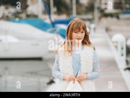 Outdoormodeporträt eines jungen jungen jungen Mädchens mit weißer Kappe, Kunstpelzweste, spielt in einem Hafen, getöntes Bild Stockfoto