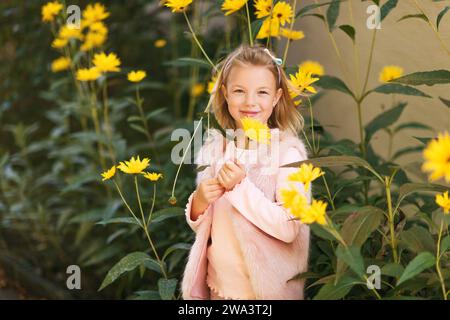 Außenporträt eines entzückenden kleinen Mädchens, das mit gelben Chrysanthemen-Blumen im Herbstgarten spielt, Kind mit hellrosa Kunstpelzjacke Stockfoto