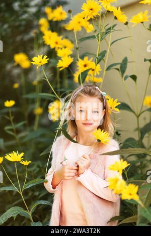 Außenporträt eines entzückenden kleinen Mädchens, das mit gelben Chrysanthemen-Blumen im Herbstgarten spielt, Kind mit hellrosa Kunstpelzjacke Stockfoto