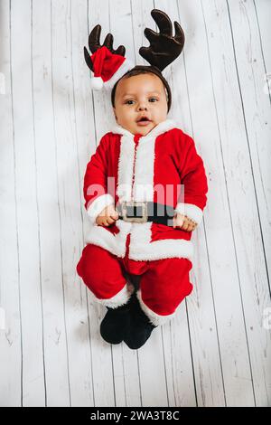 Weihnachtsporträt des entzückenden Jungen, der auf weißem Hintergrund liegt, das Weihnachtsmannskostüm und die Rentierohren trägt, Ansicht von oben Stockfoto