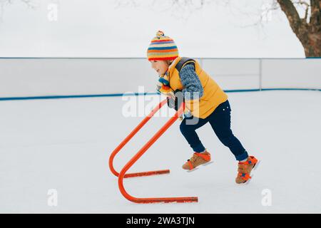 Glücklicher, lustiger kleiner Junge, der mit Unterstützung auf der Eislaufbahn trainiert, einen hellen Hut und eine gelbe Jacke trägt. Winteraktivitäten für Kinder Stockfoto