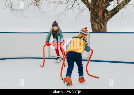 Fröhliche, lustige kleine Kinder, die mit Unterstützung auf der Eislaufbahn trainieren, helle Hüte und warme Jacken tragen. Winteraktivitäten für Kinder Stockfoto