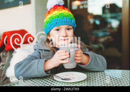 Entzückendes kleines 6-jähriges Mädchen, das heiße Schokolade im Wintercafé trinkt, große Tasse hält, grauen Mantel und bunten Hut trägt Stockfoto