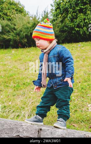 Niedlicher Kleinkind, der im Park spielt, trägt eine bunte Mütze, eine Jeansjacke und eine grüne Hose Stockfoto