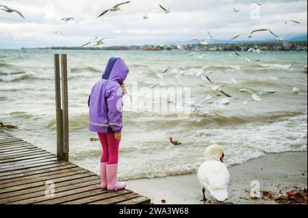 Niedliches kleines Mädchen, das bei kaltem Wetter draußen am Genfer See spielt, im Winter Vögel füttert, Rückansicht Stockfoto