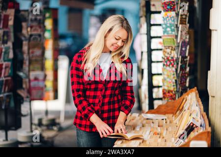 Glückliche junge blonde Leserin in einem Buchladen, die ein rotes kariertes Hemd trägt Stockfoto