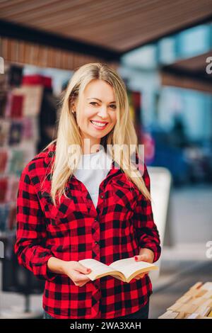 Glückliche junge blonde Leserin in einem Buchladen, die ein rotes kariertes Hemd trägt Stockfoto