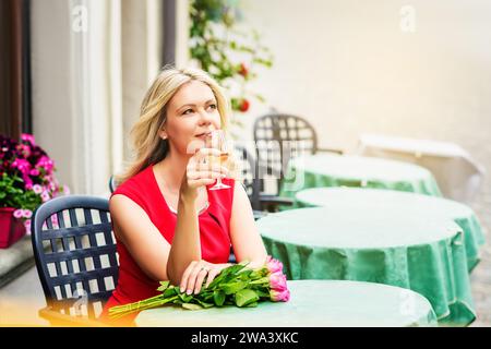 Außenporträt einer schönen blonden Frau, die einen Strauß rosa Rosen hält, im Café ruht und ein Glas Weißwein hält Stockfoto