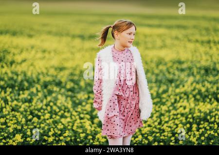 Außenporträt eines hübschen 10-jährigen Mädchens mit rosa Vintage-Kleid und Kunstfell-Bodywärmer, der auf einem Feld posiert Stockfoto