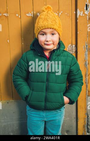 Außenporträt eines lustigen 4-5-jährigen Jungen mit grüner Winterjacke, blauer Jeans und gelbem Hut Stockfoto