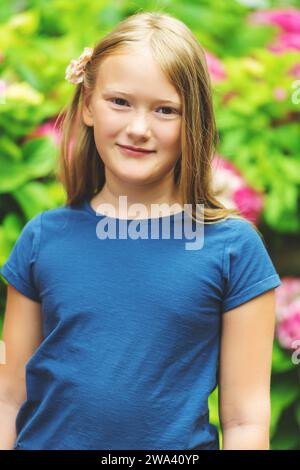 Vertikales Außenporträt eines niedlichen Mädchens von 9 Jahren im Sommergarten, gestützt auf einen Zaun, mit blauem T-Shirt Stockfoto