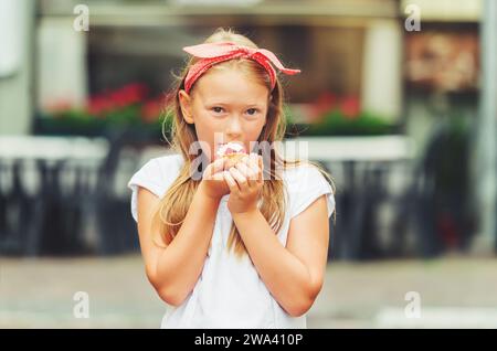 Lustiges kleines Mädchen, das draußen Kuchen isst, mit rotem Punktband Stockfoto