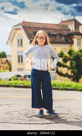 Lustiges Outdoor-Porträt eines süßen kleinen 9-10-jährigen Mädchens mit blauem Oberteil, Jeans-Culottes und Brille Stockfoto