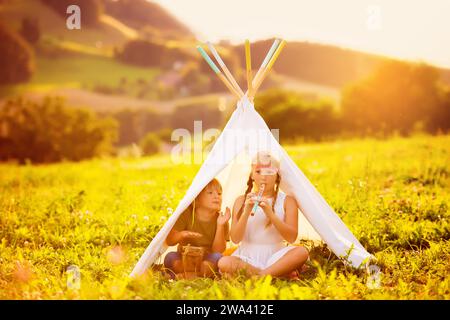 Zwei glückliche Kinder sitzen in einem Zelt an einem schönen warmen Abend, spielen Flöte und Trommelmusik. Bruder und Schwester haben viel Spaß miteinander Stockfoto