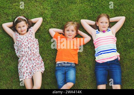 Glückliche Kinder, die sich im Freien amüsieren. Kinder spielen im Sommerpark. Kleiner Junge und zwei Mädchen liegen auf grünem, frischem Gras Stockfoto