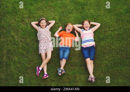 Glückliche Kinder, die sich im Freien amüsieren. Kinder spielen im Sommerpark. Kleiner Junge und zwei Mädchen liegen auf grünem, frischem Gras Stockfoto