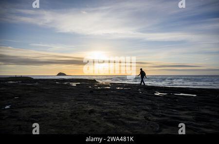 Mann, der bei Sonnenuntergang am Strand von Muriwai auf den Felsen läuft. Oaia Island in der Ferne. Auckland. Stockfoto