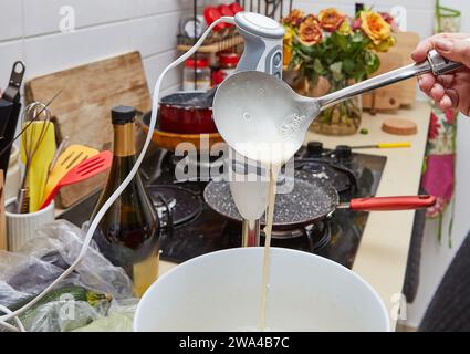 Nahaufnahme einer Frau, die in einer gemütlichen rustikalen Küche steht und hausgemachte Pfannkuchenmischung aus einer Kelle in eine Schüssel gießt. Die Mischung taucht aus und schafft so ein entzückendes Stockfoto