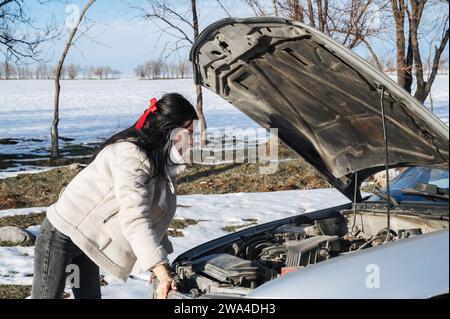 Mädchen schaut unter der Motorhaube auf den Motor eines kaputten Autos auf der Straße im Winter Stockfoto