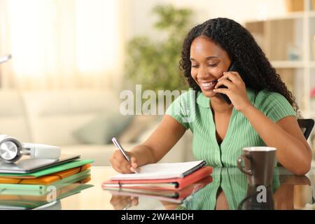 Schwarzer glücklicher Schüler, der am Telefon anruft, während er studiert und sich zu Hause Notizen macht Stockfoto
