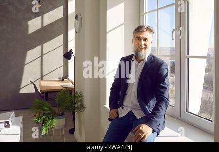 Reifer Geschäftsmann im Anzug sitzt am Fenster im Büro, blickt in die Kamera und lächelt. Stockfoto