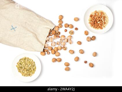 Große Tüte Sackleinen mit Walnüssen und Haselnüssen. Nüsse auf weißem Hintergrund verstreut. Auf dem Brett befinden sich zwei weiße Teller mit Mandeln und Erdnüssen, Pumpe Stockfoto