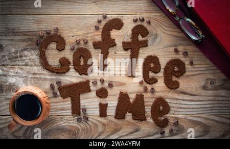 Kaffeezeit geschrieben von gemahlenem Kaffee auf hölzernem Hintergrund. Tasse heißer Kaffee mit Dampf. Stockfoto