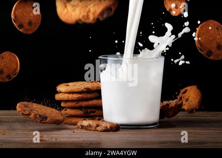 Ein Glas Milch mit leckeren Keksen. Milch wird mit Spritzern in eine Tasse gegeben. Kekse fliegen in der Luft und fallen mit Krümeln auf den Tisch. Schwarzer Hintergrund Stockfoto