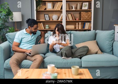 Fröhliches, gemischtes Millennial-Paar, das Zeit mit Laptop und Buch zu Hause verbringt, glückliche junge Interracial-Liebhaber sitzen auf Sofa mit Computer, freiberuflich mit Stockfoto
