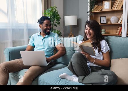 Fröhliches, gemischtes Millennial-Paar, das Zeit mit Laptop und Buch zu Hause verbringt, glückliche junge Interracial-Liebhaber sitzen auf Sofa mit Computer, freiberuflich mit Stockfoto